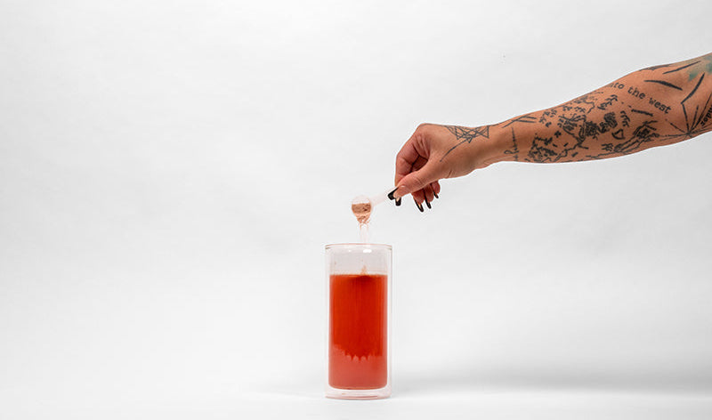 A tattooed hand holding a small dropper over a clear glass container filled with a red liquid, photographed against a seamless white studio background.